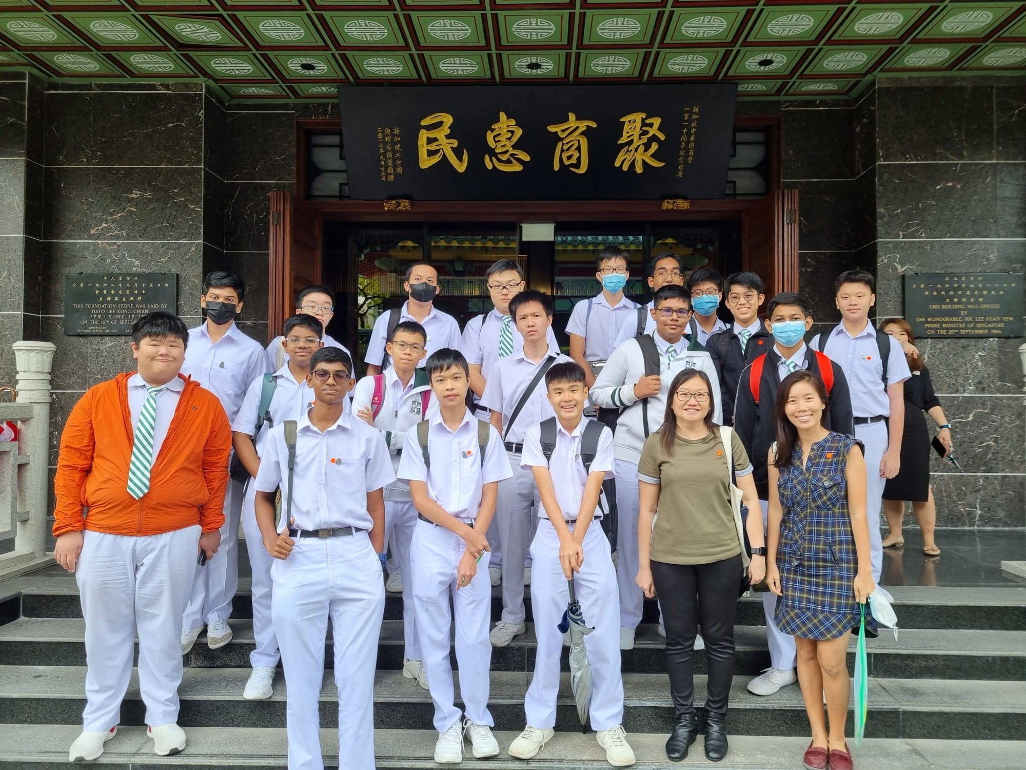 A group of students in white uniforms with two adults posing on steps outside a building with Chinese characters above the entrance.